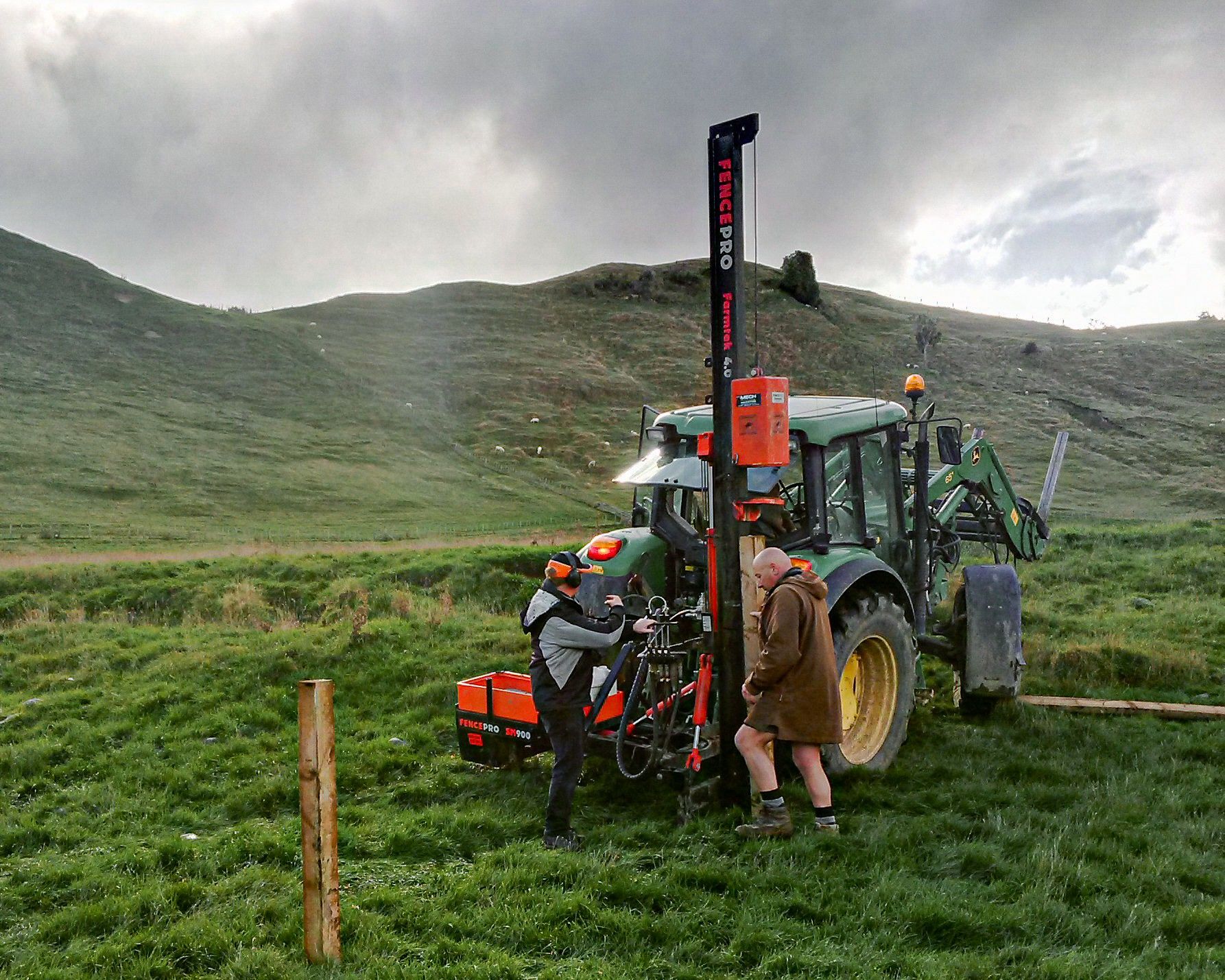 Farmers operating a FencePro Farmtek post driver mounted on a tractor during fencing in rural New Zealand.